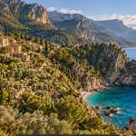 Kamnita vasica nad turkiznim zalivom na Mallorci, z gorami Serra de Tramuntana v ozadju.