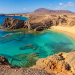 Peščena plaža s turkiznim morjem in vulkansko pokrajino na Lanzarotu.